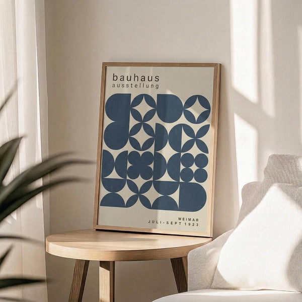 May include: A framed Bauhaus exhibition poster with geometric blue circles and shapes on cream background, displayed on a round wooden side table next to a beige upholstered chair. Natural sunlight streams across the minimalist room setting, creating soft shadows. The poster features vintage typography and is housed in a thin wooden frame.