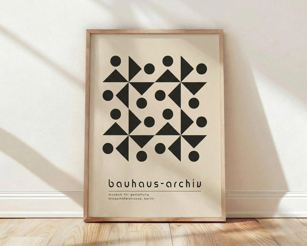 May include: Framed geometric poster with black triangular and circular shapes arranged in symmetrical pattern on cream background, displayed against white wall on light wooden floor with natural shadows