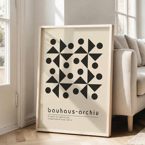 May include: Framed geometric art print with black triangles and circles arranged in symmetrical pattern on cream background, displayed in bright living room corner next to beige upholstered chair. Natural light streams through white-framed windows onto light wood floors. The minimalist poster features bold Bauhaus-style shapes with museum text below the geometric design.