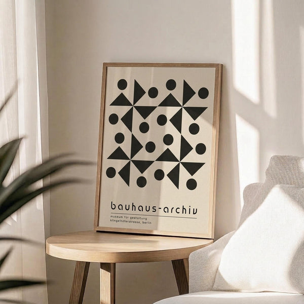 May include: Framed geometric poster with black triangles and circles in repeating pattern on cream background, displayed on round wooden stool next to beige upholstered chair. Sunlight casts shadows across neutral-toned room with plant leaves visible. Poster features Bauhaus-style design with museum text at bottom in thin wooden frame.