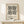 May include: A framed geometric art print with black triangles and circles arranged in a symmetrical pattern on cream background, displayed on a light wood shelf beside a small wrapped gift with evergreen sprig. The thin wooden frame leans against a white wall in soft natural lighting.