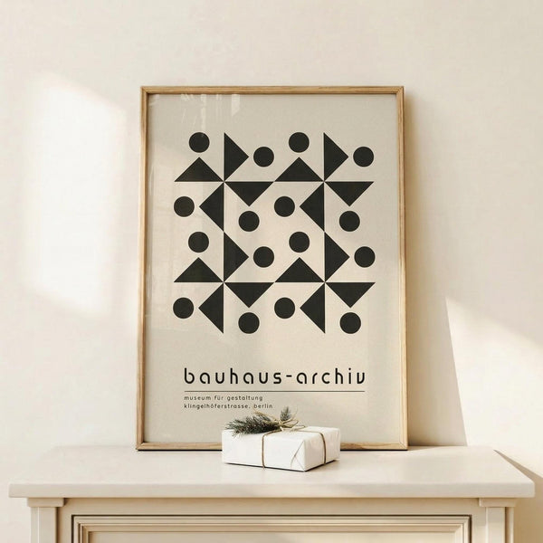 May include: A framed geometric art print with black triangles and circles arranged in a symmetrical pattern on cream background, displayed on a light wood shelf beside a small wrapped gift with evergreen sprig. The thin wooden frame leans against a white wall in soft natural lighting.