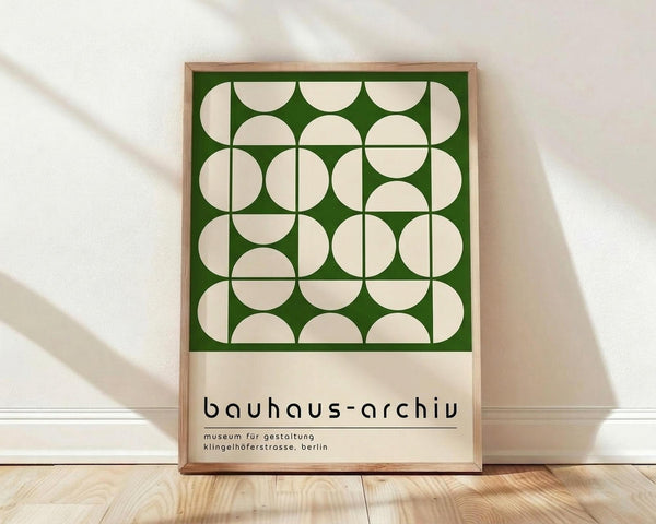 May include: Framed geometric poster with green background and cream-colored overlapping circles arranged in a grid pattern, displayed against white wall on light wood floor. Natural shadows cast across the scene. Text reads "bauhaus-archiv" and "museum für gestaltung klingelhöferstrasse, berlin" below the design. Light wooden frame surrounds the minimalist mid-century modern artwork.