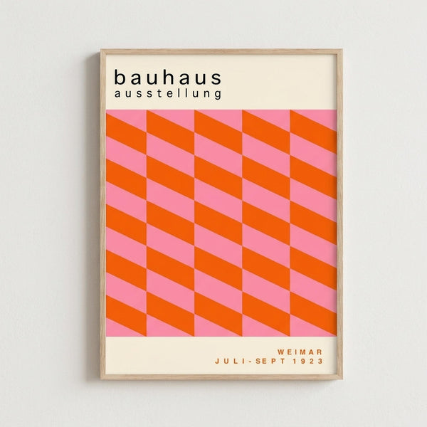 Framed vintage-style exhibition poster with geometric pattern of orange and pink interlocking cubes creating an optical illusion effect. Black text at top reads "bauhaus ausstellung" and bottom shows "WEIMAR JULI-SEPT 1923". The artwork features bold modernist design with alternating bright orange and soft pink parallelograms arranged in repeating rows. Natural wood frame against white wall background.