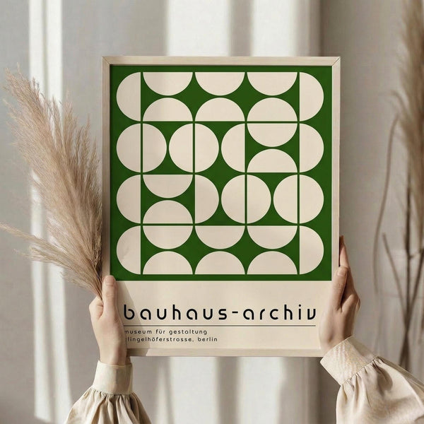 May include: Person holding framed geometric poster with green background and cream-colored interlocking circles pattern. Light wood frame, minimalist museum-style design. Soft neutral interior setting with dried pampas grass and flowing curtains. Hands wearing cream-colored clothing with gathered sleeves.