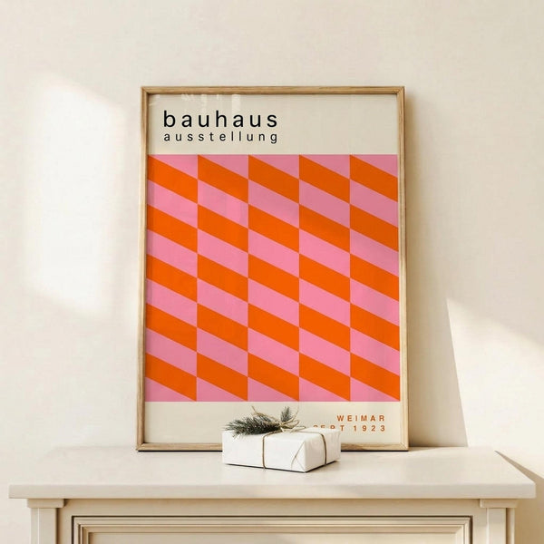 May include: Framed Bauhaus exhibition poster with geometric orange and pink cube pattern displayed on white wooden shelf. Light wood frame contains vintage-style design with "bauhaus ausstellung" text and "Weimar 1923" at bottom. Small white wrapped gift with evergreen sprig sits beside frame against cream wall.