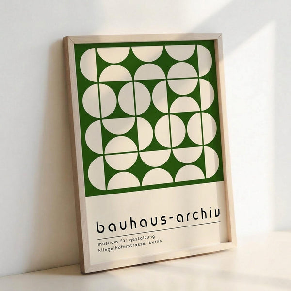Framed poster featuring geometric pattern of white circles on forest green background with black text reading "bauhaus-archiv" and "museum für gestaltung klingelhofstrasse, berlin" below. Light wood frame leaning against white wall, casting subtle shadow.