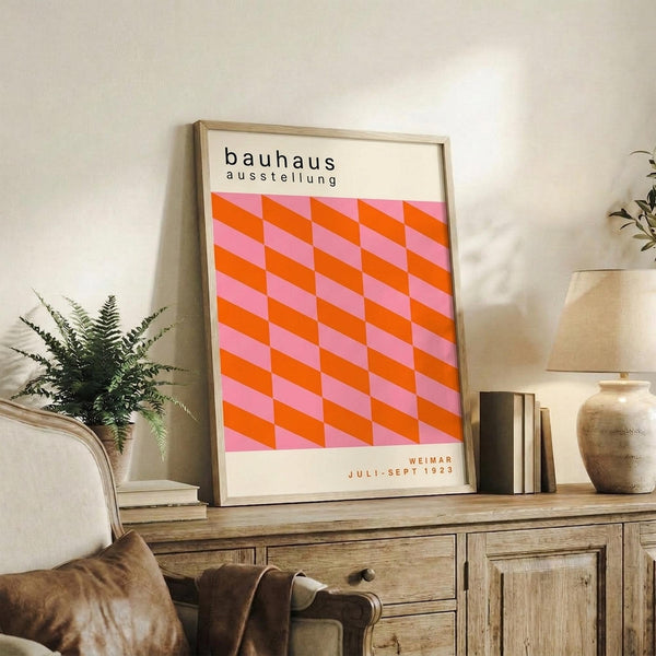 May include: Framed Bauhaus exhibition poster with geometric orange and pink cube pattern displayed on rustic wood sideboard in modern living room setting, surrounded by fern plant, ceramic vase, stacked books, and beige upholstered chair with leather cushion