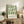 May include: Framed green and cream geometric poster with Bauhaus-inspired circular pattern displayed on rustic wood sideboard next to fern plant, books, and ceramic vase in modern living room with beige chair and neutral decor