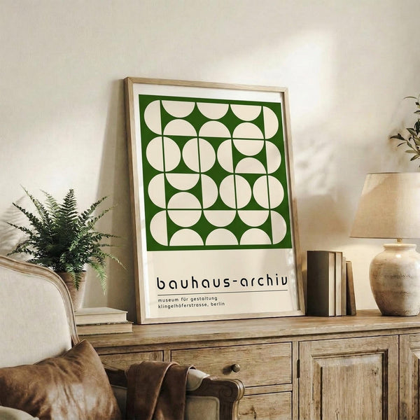 May include: Framed green and cream geometric poster with Bauhaus-inspired circular pattern displayed on rustic wood sideboard next to fern plant, books, and ceramic vase in modern living room with beige chair and neutral decor