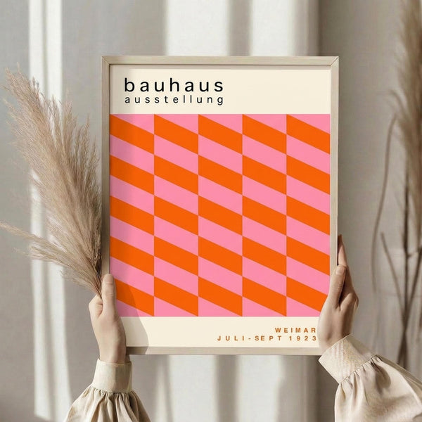 May include: Person in beige sweater holding framed geometric poster with pink and orange diamond cube pattern. Black text reads "bauhaus ausstellung" at top and "WEIMAR JULI-SEPT 1923" at bottom. Light wood frame, neutral background with dried wheat stems visible on left side.