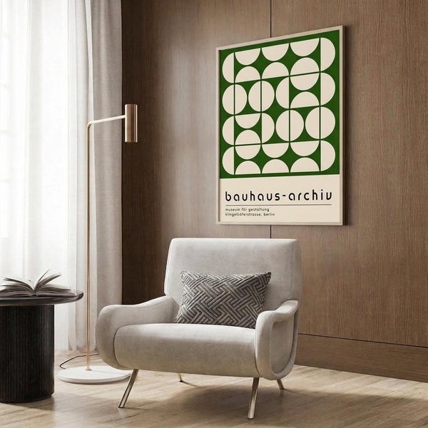 May include: Framed Bauhaus museum poster with green and cream geometric circle pattern displayed on wood-paneled wall above beige upholstered chair with patterned throw pillow. Modern brass floor lamp and dark round side table with open book visible. Neutral-toned contemporary interior setting with natural lighting from window.
