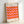 May include: Framed Bauhaus exhibition poster with geometric pattern of alternating orange and pink parallelograms creating a three-dimensional cube effect. White background with black text reading "bauhaus ausstellung" at top and "WEIMAR JULI-SEPT 1923" at bottom. Light wood frame leaning against white wall on white surface, casting soft shadow.