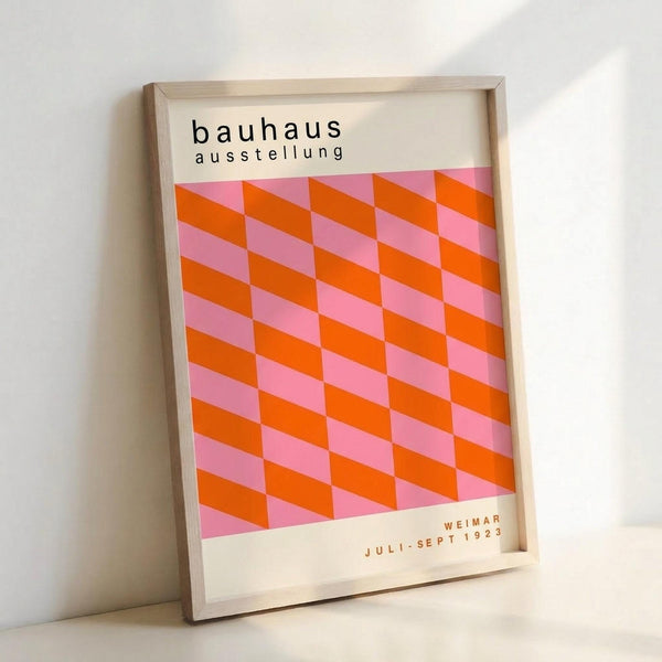 May include: Framed Bauhaus exhibition poster with geometric pattern of alternating orange and pink parallelograms creating a three-dimensional cube effect. White background with black text reading "bauhaus ausstellung" at top and "WEIMAR JULI-SEPT 1923" at bottom. Light wood frame leaning against white wall on white surface, casting soft shadow.