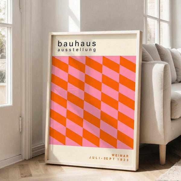 May include: Framed poster with geometric pink and orange cube pattern leaning against white wall next to beige sofa. Print features "bauhaus ausstellung" text at top and "WEIMAR JULI-SEPT 1923" at bottom on cream background. Light wood frame, bright natural lighting from nearby windows, hardwood floors with area rug visible.