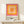 May include: Framed geometric art print featuring concentric squares in warm tones - pink outer square, orange middle, peach and coral inner squares. Text reads "bauhaus-archiv museum für gestaltung" below the colorful design. Displayed on light wood shelf against cream wall with small wrapped gift beside the thin gold frame.