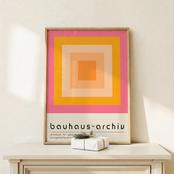 May include: Framed geometric art print featuring concentric squares in warm tones - pink outer square, orange middle, peach and coral inner squares. Text reads "bauhaus-archiv museum für gestaltung" below the colorful design. Displayed on light wood shelf against cream wall with small wrapped gift beside the thin gold frame.