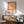 May include: Framed Bauhaus-style poster with concentric squares in orange, pink, and peach tones displayed above a mid-century wooden credenza. The geometric artwork features warm gradient colors transitioning from deep orange center to pink border. Setting shows modern interior with sage green upholstered chair, white ceramic lamp, and natural sunlight casting shadows on white wall.