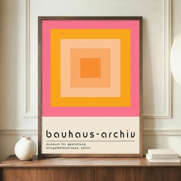 May include: Framed geometric poster with concentric squares in pink, orange, peach and coral tones displayed on wooden sideboard next to white ceramic vase and books. The minimalist artwork features nested rectangles creating an ombré effect from bright pink outer border to deep orange center square. Black text below reads "bauhaus-archiv" and mentions Berlin museum details. Warm interior setting with cream walls and paneled wainscoting.