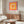 May include: Colorful geometric poster with concentric squares in pink, orange, yellow and peach tones hanging above a wooden sideboard. Modern living room setting features cream upholstered chair, brass floor lamp, glass-top coffee table, and neutral walls. Poster shows museum exhibition text at bottom.