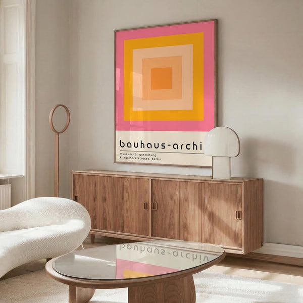 May include: Colorful geometric poster with concentric squares in pink, orange, yellow and peach tones hanging above a wooden sideboard. Modern living room setting features cream upholstered chair, brass floor lamp, glass-top coffee table, and neutral walls. Poster shows museum exhibition text at bottom.