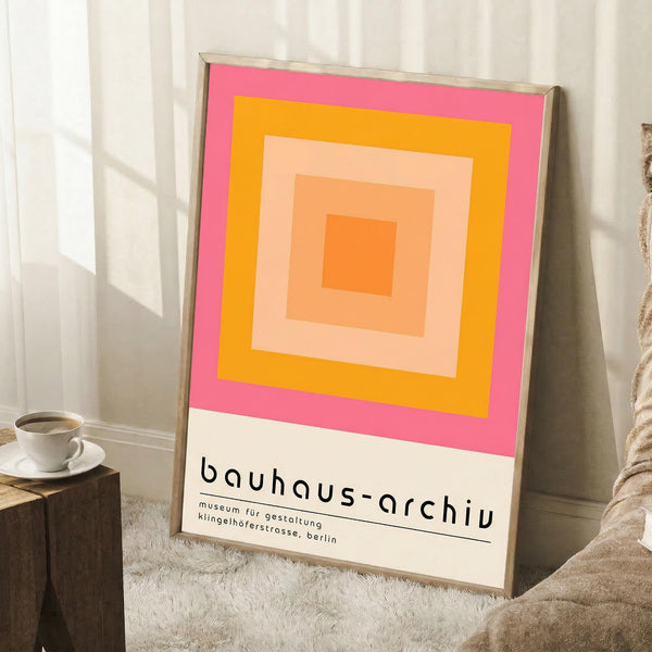 May include: A framed poster with concentric squares in pink, orange, yellow and peach tones displaying Bauhaus-Archiv museum information at bottom, leaning against white paneled wall on textured cream rug next to wooden block with white coffee cup and beige knit throw blanket in bright natural lighting.