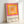 Framed poster with concentric squares in vibrant pink, yellow, orange, and peach tones creating a geometric gradient pattern. Text at bottom reads "bauhaus-archiv" and "museum für gestaltung klingelhöferstrasse, berlin" in lowercase black letters. Light wooden frame against white wall, casting subtle shadow.