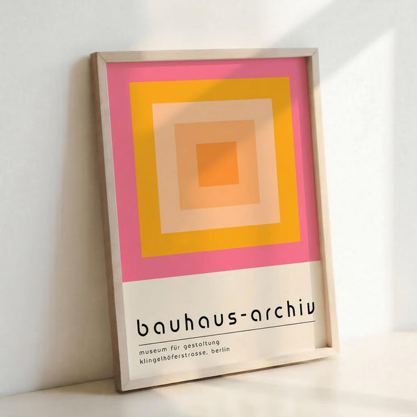 Framed poster with concentric squares in vibrant pink, yellow, orange, and peach tones creating a geometric gradient pattern. Text at bottom reads "bauhaus-archiv" and "museum für gestaltung klingelhöferstrasse, berlin" in lowercase black letters. Light wooden frame against white wall, casting subtle shadow.