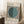 May include: Framed Bauhaus exhibition poster featuring black and white vertical stripes with a large teal circle overlay, displayed in light wooden frame leaning against white wall corner. Coffee cup on wooden block and beige textured throw blanket visible nearby on white fluffy rug.