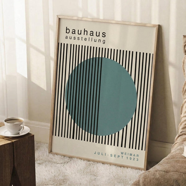 May include: Framed Bauhaus exhibition poster featuring black and white vertical stripes with a large teal circle overlay, displayed in light wooden frame leaning against white wall corner. Coffee cup on wooden block and beige textured throw blanket visible nearby on white fluffy rug.