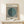 May include: Framed Bauhaus exhibition poster with black and white vertical stripes and teal green circle, displayed on light wood console table next to small white wrapped gift with natural twine and evergreen sprig, against cream wall with soft natural lighting