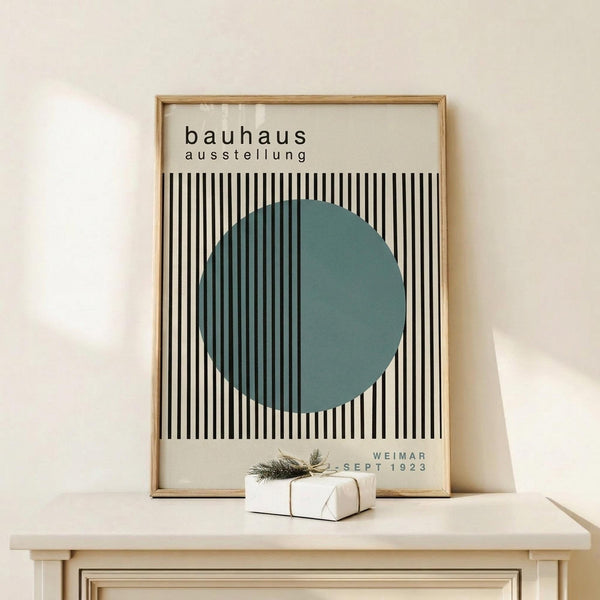 May include: Framed Bauhaus exhibition poster with black and white vertical stripes and teal green circle, displayed on light wood console table next to small white wrapped gift with natural twine and evergreen sprig, against cream wall with soft natural lighting