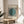May include: Minimalist Bauhaus exhibition poster print in light wood frame displayed on round wooden side table. Features teal circle overlapping black and white vertical stripes with German text. Sunlit modern living room with beige chair, plant shadows, and cream walls creates serene Scandinavian-style interior setting.