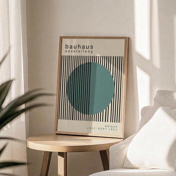 May include: Minimalist Bauhaus exhibition poster print in light wood frame displayed on round wooden side table. Features teal circle overlapping black and white vertical stripes with German text. Sunlit modern living room with beige chair, plant shadows, and cream walls creates serene Scandinavian-style interior setting.