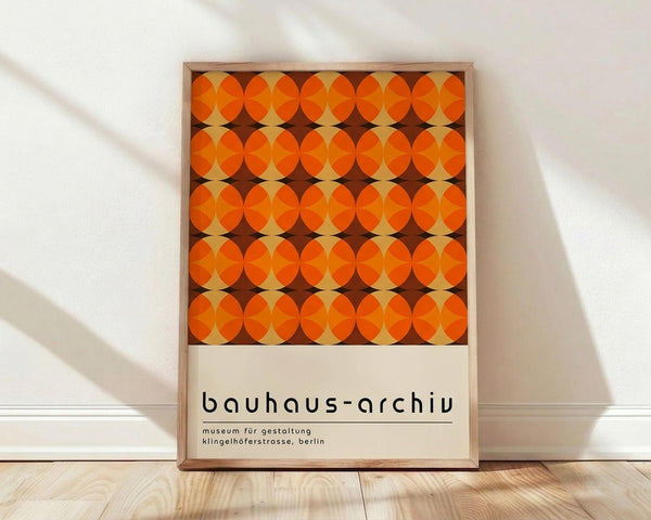 May include: Framed poster with geometric orange and brown circular pattern in Bauhaus style, displayed against white wall on light wooden floor. Design features repeating overlapping circles creating diamond shapes between them. Simple wooden frame with museum text at bottom on cream background.