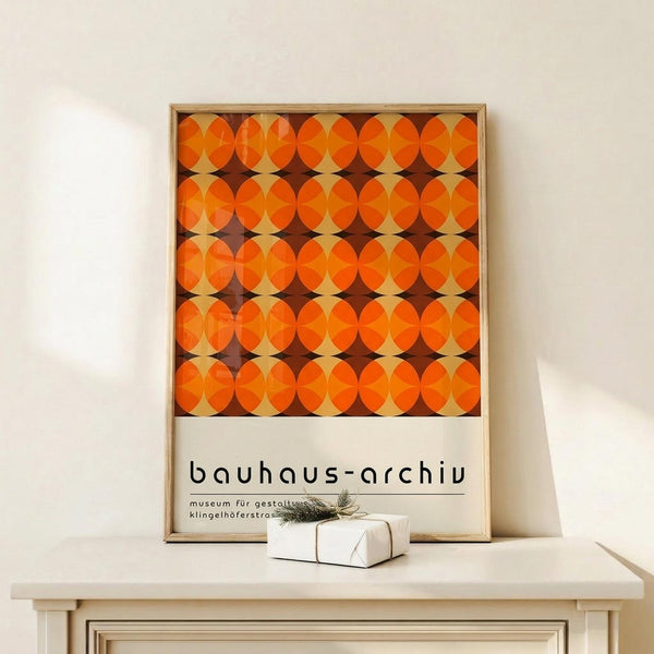 May include: Framed poster with geometric orange and brown circular pattern displayed on light wood surface against white wall. Small wrapped gift with twine and evergreen sprig sits in front of wooden frame. Poster features repeating overlapping circles creating retro mid-century modern design in warm autumn tones with museum text below pattern.