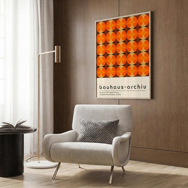 May include: Bauhaus-style poster with orange circular pattern mounted on wood-paneled wall above modern beige armchair with geometric patterned pillow. Scene shows contemporary living room with brass floor lamp, dark round side table with open book, and neutral curtains. Poster features repetitive orange circles on brown background in mid-century modern aesthetic.