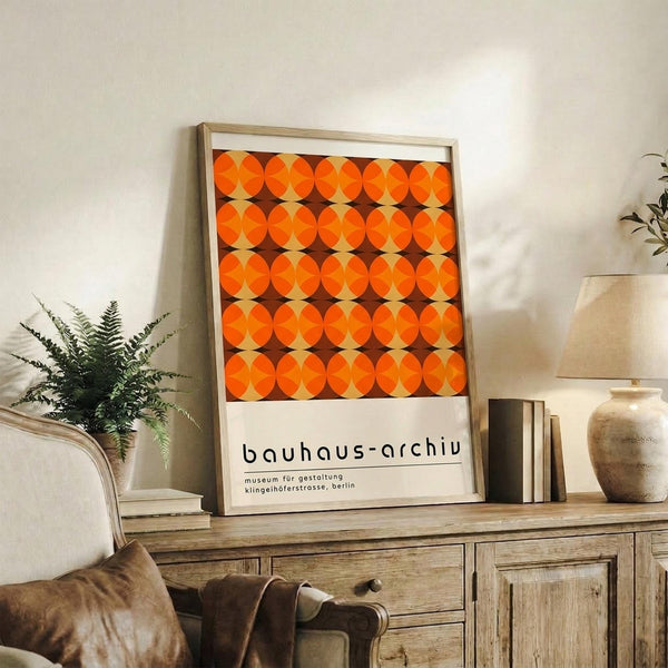 May include: Framed Bauhaus-style poster with geometric pattern of orange and brown overlapping circles displayed on rustic wooden sideboard alongside fern plant, ceramic vases, and books in warm-toned living room setting with beige armchair visible