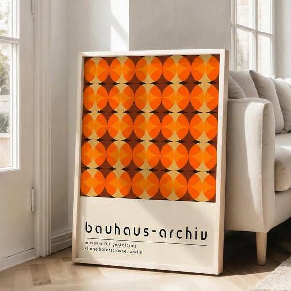 May include: Framed poster with orange and brown geometric circle pattern leaning against white wall next to beige upholstered chair. The artwork features overlapping circles creating a retro mid-century modern design in warm orange tones with dark brown accents. White frame with museum text at bottom. Bright room with large windows and wooden floors.