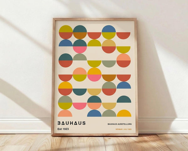 May include: Framed geometric art print leaning against white wall on light wood floor. Design features colorful semicircles in rows - blues, oranges, yellows, pinks, and greens on cream background. Text reads "BAUHAUS Est 1923" and "BAUHAUS AUSSTELLUNG WEIMAR JULI 1923" at bottom. Natural wood frame, soft window light casting diagonal shadows across textured white wall.