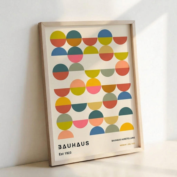 A framed poster featuring a geometric pattern of colorful circles arranged in horizontal rows. Each circle is divided in half with different color combinations including blues, greens, oranges, yellows, and pinks against a cream background. The design has a mid-century modern aesthetic with "BAUHAUS Est 1923" and "BAUHAUS AUSSTELLUNG WEIMAR | JULI 1923" text at the bottom. The poster is displayed in a light wood frame leaning against a white wall.