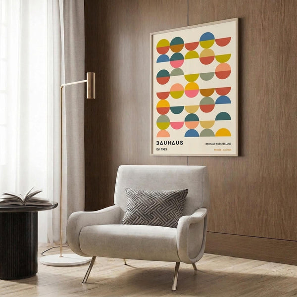 May include: Framed Bauhaus poster with colorful geometric circles in rows, displayed on wood paneled wall above beige upholstered chair with patterned pillow. Modern brass floor lamp and round black side table with open book nearby. Neutral tones with warm wood textures throughout contemporary living space.