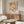 May include: Framed Bauhaus poster with colorful geometric half-circles in rows, displayed above a mid-century modern wood credenza in minimalist living room. White upholstered chair, glass coffee table, and floor lamp visible. Poster features vibrant blues, oranges, greens, and yellows in systematic pattern against cream background.