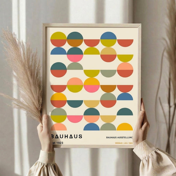 May include: Person holding framed Bauhaus-style poster featuring geometric pattern of colorful half-circles in rows. Circles show vibrant colors including yellow, orange, coral, teal, blue, and sage green on cream background. Light wood frame, neutral interior setting with dried pampas grass visible. Hands wearing beige long-sleeved top with gathered cuffs.