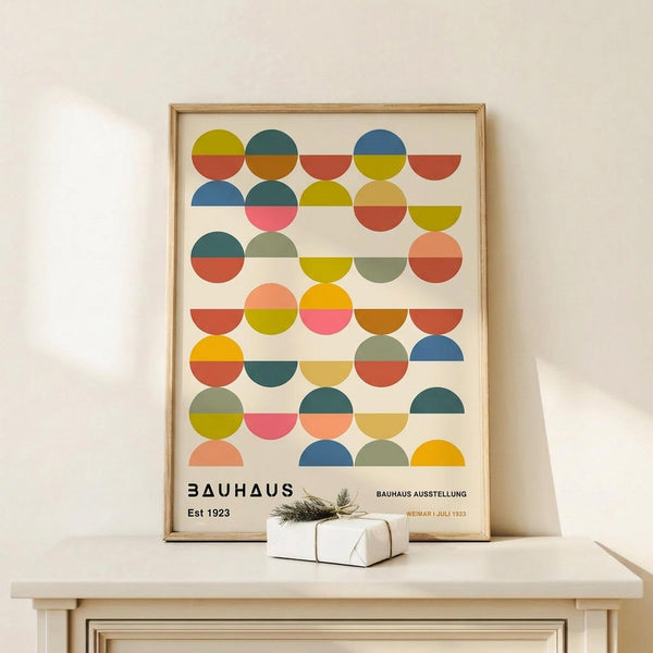May include: Framed Bauhaus-style poster with colorful geometric circles in rows, featuring split-tone designs in blues, greens, yellows, oranges, and pinks on cream background. Natural wood frame sits on light wooden console table beside small wrapped gift with twine and pine sprig against neutral wall.