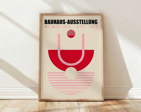 May include: Framed geometric poster with cream background featuring red and pink abstract shapes - a red semi-circle with pink handle-like curves above, striped red circle at top, and striped pink semi-circle below. Black text reads "BAUHAUS-AUSSTELLUNG" and "WEIMAR JULI-SEPT 1923". Natural wood frame leaning against white wall on light wooden floor with soft window shadows.