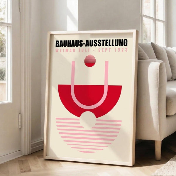 May include: A framed poster with cream background featuring bold red and pink geometric shapes including a striped circle, semicircle, and triangle arrangement. Black text reads "BAUHAUS-AUSSTELLUNG" and "WEIMAR JULI-SEPT 1923" at top. The light wood frame leans against white walls near a beige upholstered sofa with wooden legs on light hardwood flooring in a bright, minimalist living room with large windows.