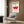 May include: Geometric Bauhaus exhibition poster in cream with bold red and pink shapes mounted on wood-paneled wall above beige upholstered chair with patterned pillow, brass floor lamp, and dark side table with open book in modern living room setting