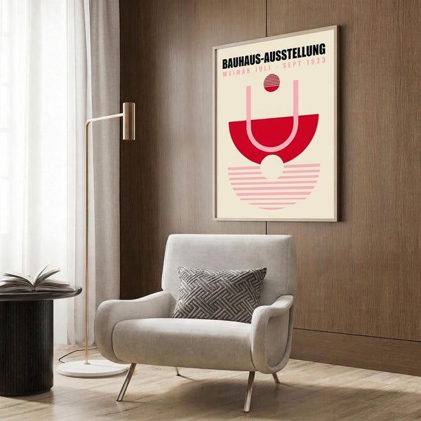 May include: Geometric Bauhaus exhibition poster in cream with bold red and pink shapes mounted on wood-paneled wall above beige upholstered chair with patterned pillow, brass floor lamp, and dark side table with open book in modern living room setting