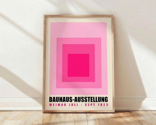 May include: Framed vintage-style poster with concentric pink squares in graduated shades from light to bright magenta, featuring black text reading "BAUHAUS-AUSSTELLUNG WEIMAR JULI - SEPT 1923" at bottom, displayed in light wood frame against white textured wall on wooden floor with soft natural lighting casting diagonal shadows